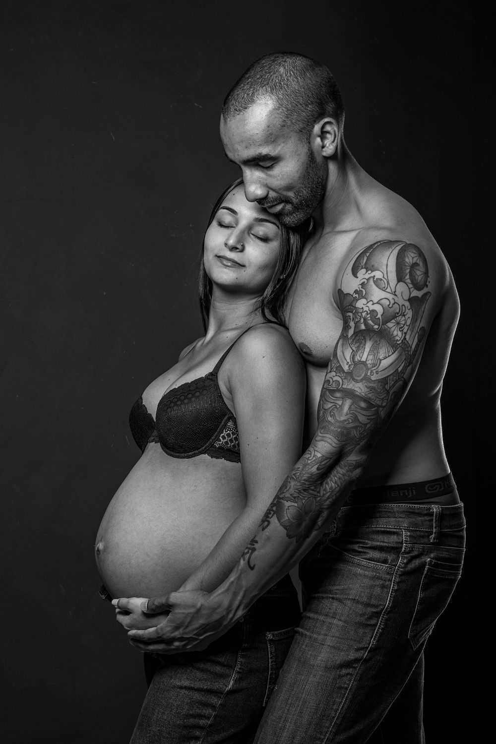 Intimate black and white maternity photo showing expecting couple embracing, with visible tattoo on man's arm.