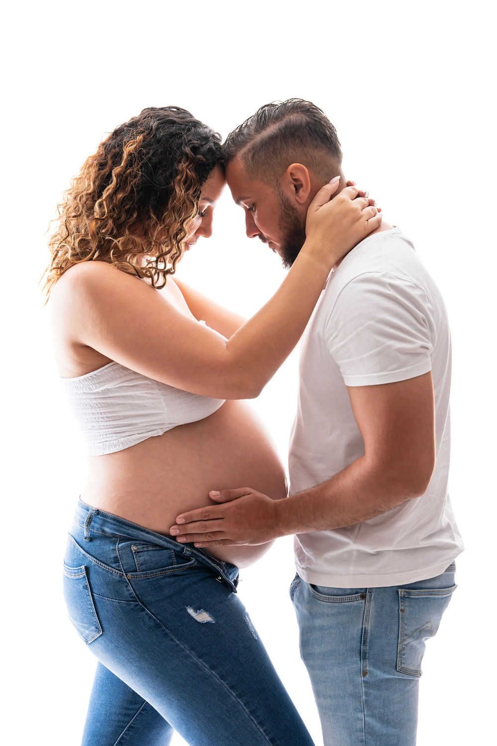 A tender maternity photo shows an expecting couple embracing in white shirts and jeans against a light background.