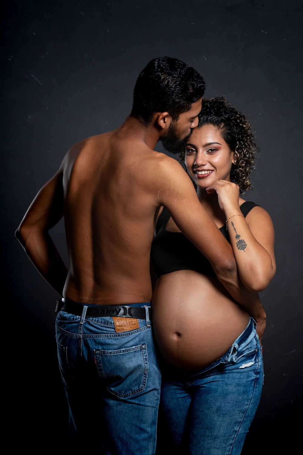 Expecting couple in denim jeans poses intimately against dark background during pregnancy photoshoot.