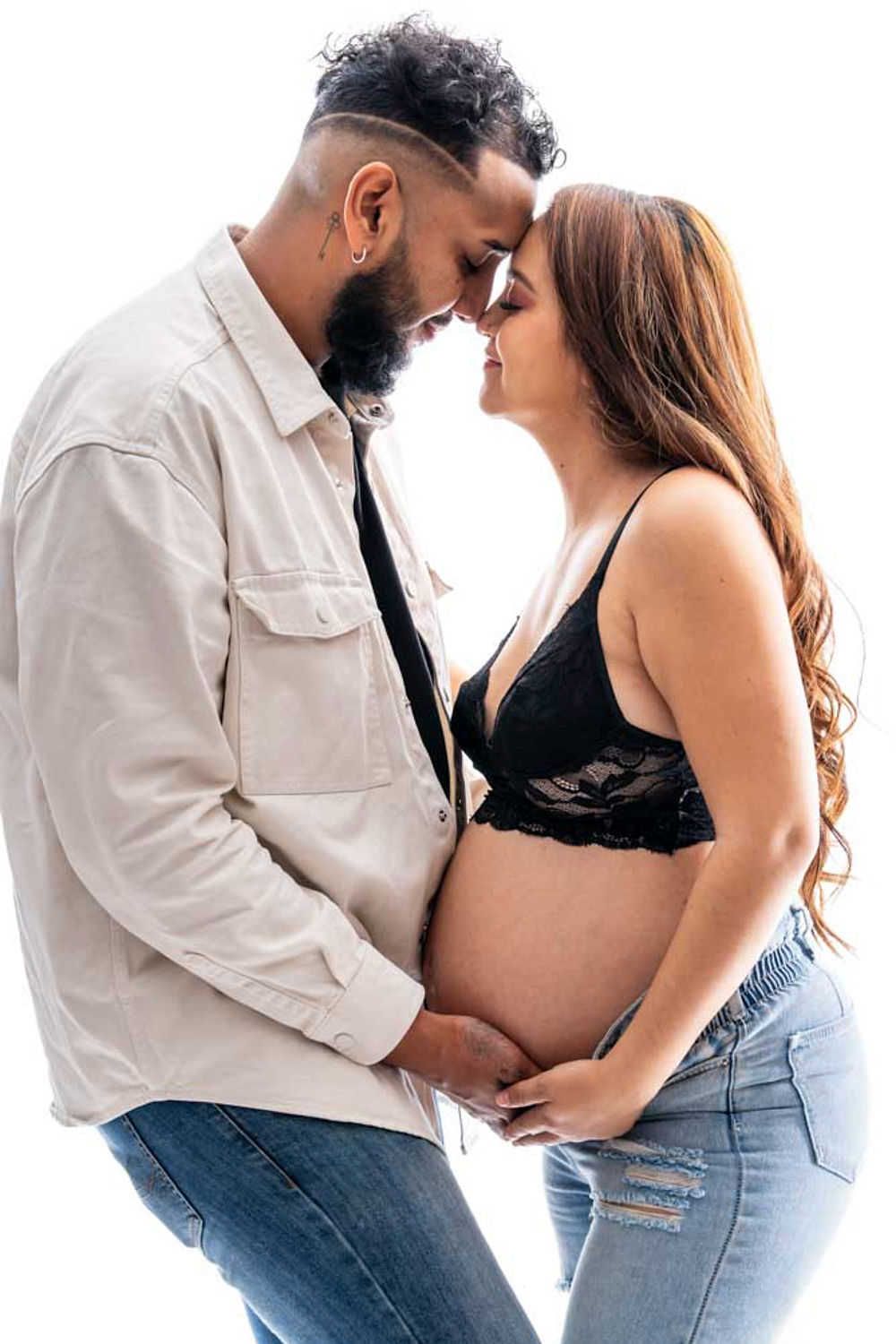 A loving couple shares an intimate moment during maternity photoshoot, dressed in casual attire against white backdrop.