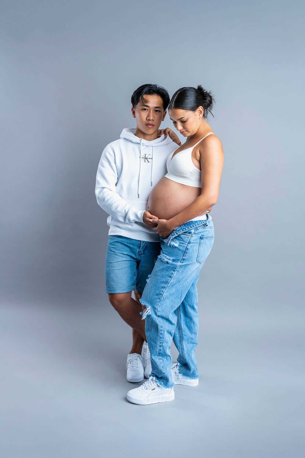 An intimate maternity portrait of a couple in casual attire posing together against a grey backdrop.