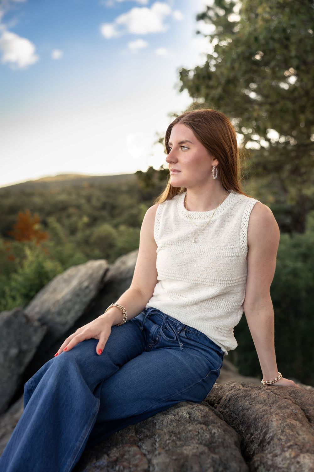 A person in a white sleeveless top and blue jeans reclines thoughtfully on large rocks against a scenic mountain backdrop.