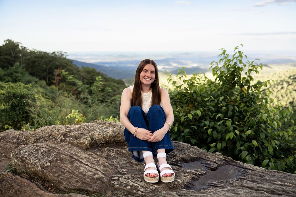 A scenic mountain overlook features someone sitting on a rocky outcrop with rolling hills and greenery in the background.