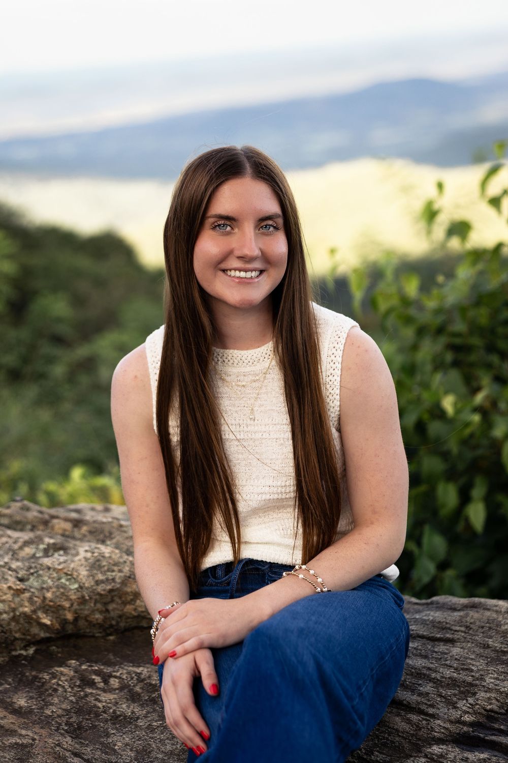 Person in white sleeveless top and blue jeans sits on rocky ledge against scenic mountain landscape at sunset.