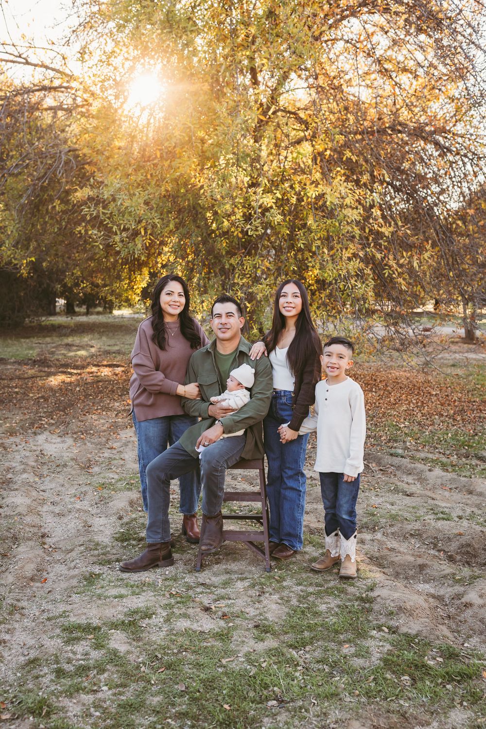 A family sits together on a wooden bench in a park during golden hour with autumn trees in the background.