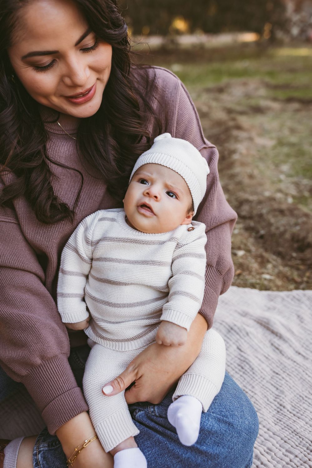 A baby in a white knit outfit sits on a blanket outdoors during an autumn photoshoot.