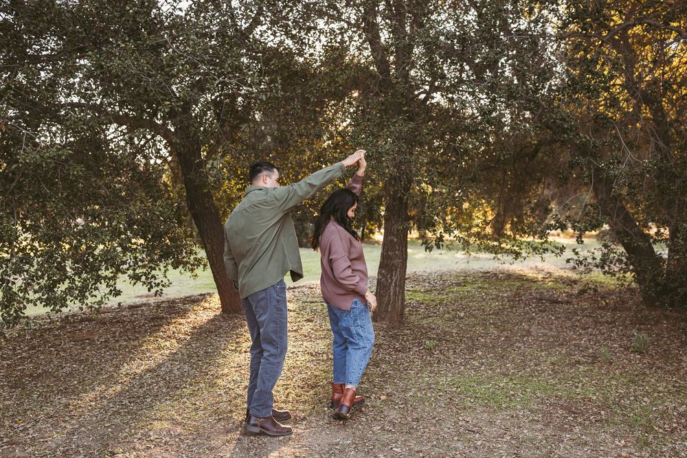 A couple walking together through a wooded area at sunset, with one person raising their arm playfully.