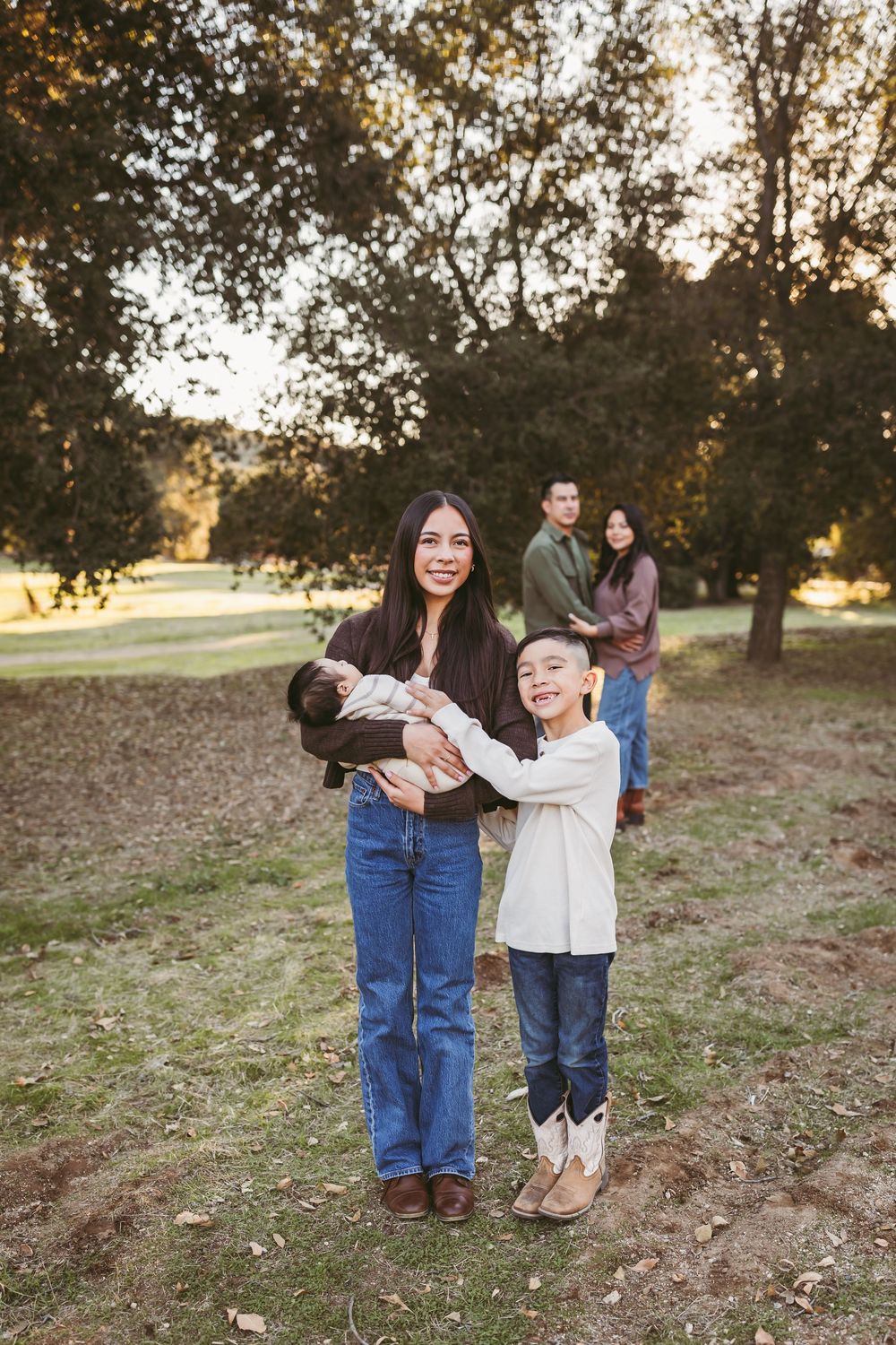 A family enjoying a sunset photoshoot in a park with trees, with a young child reaching up to a baby.