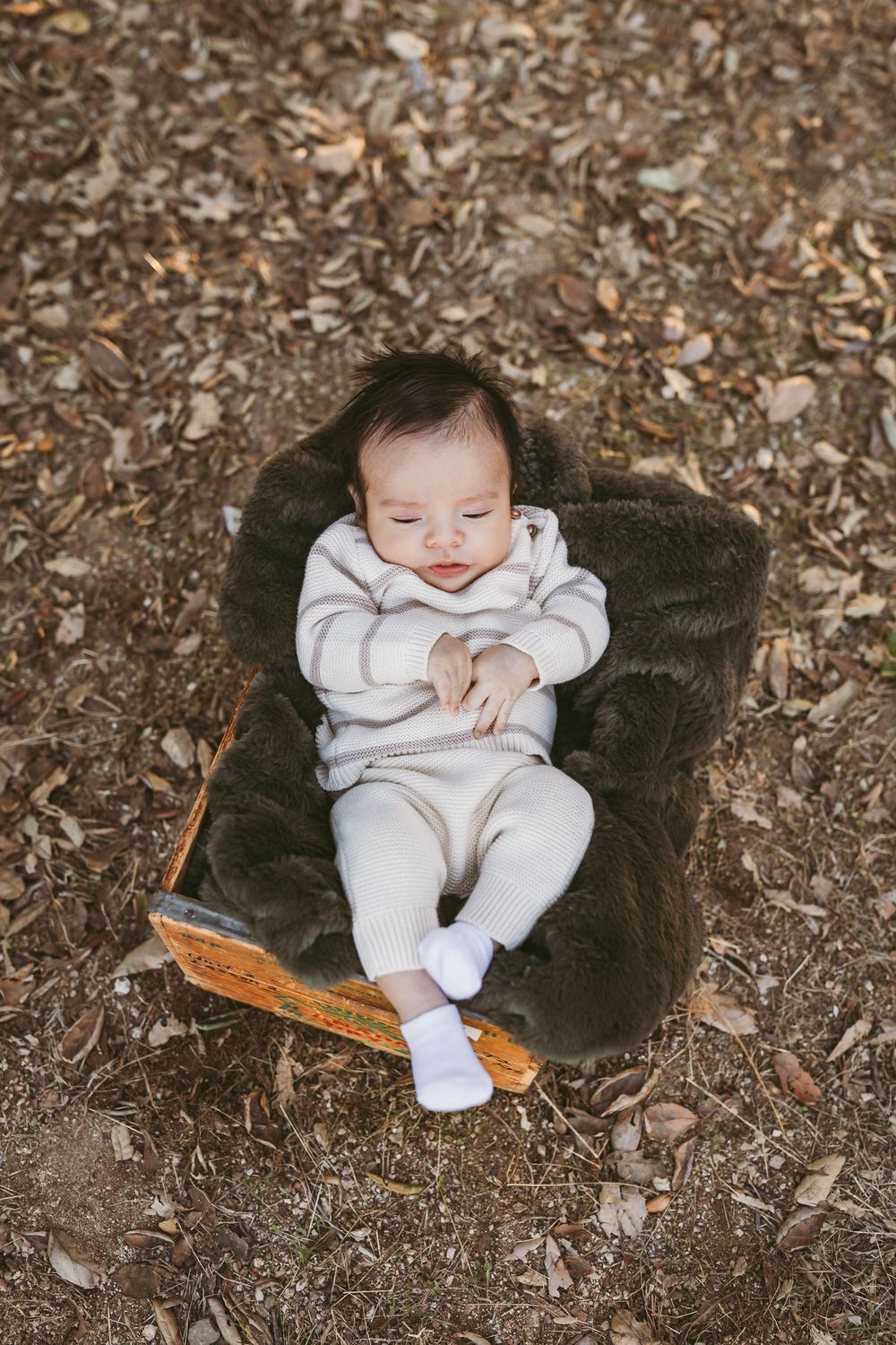 Baby in white outfit rests peacefully on dark blanket surrounded by autumn leaves on ground.