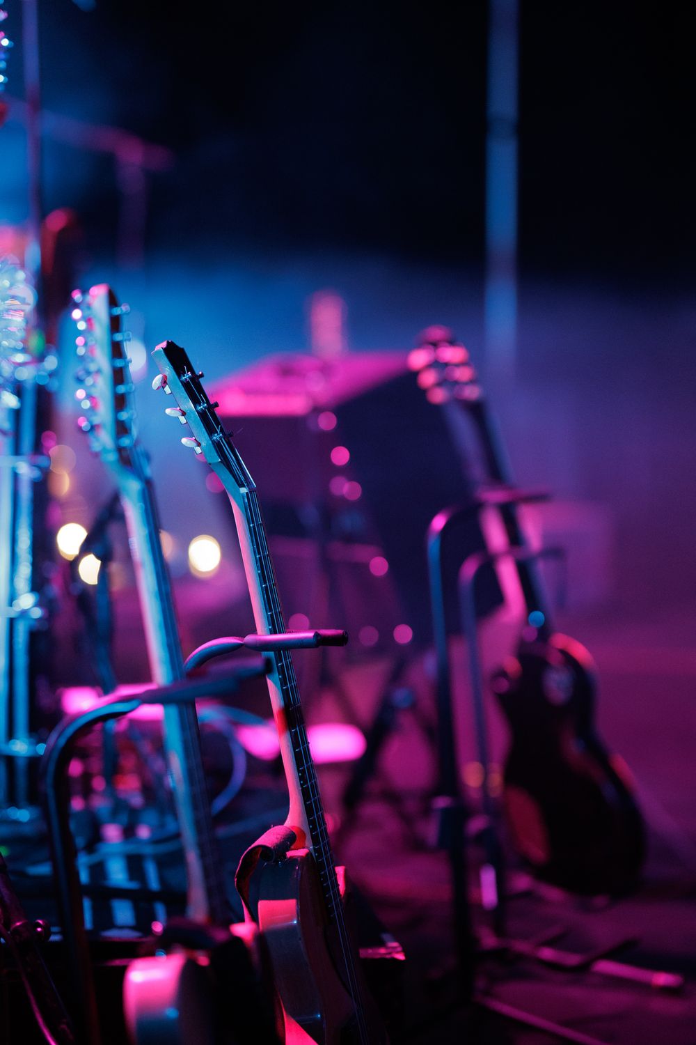 Electric guitars on stands await performers on stage bathed in blue and pink concert lighting.