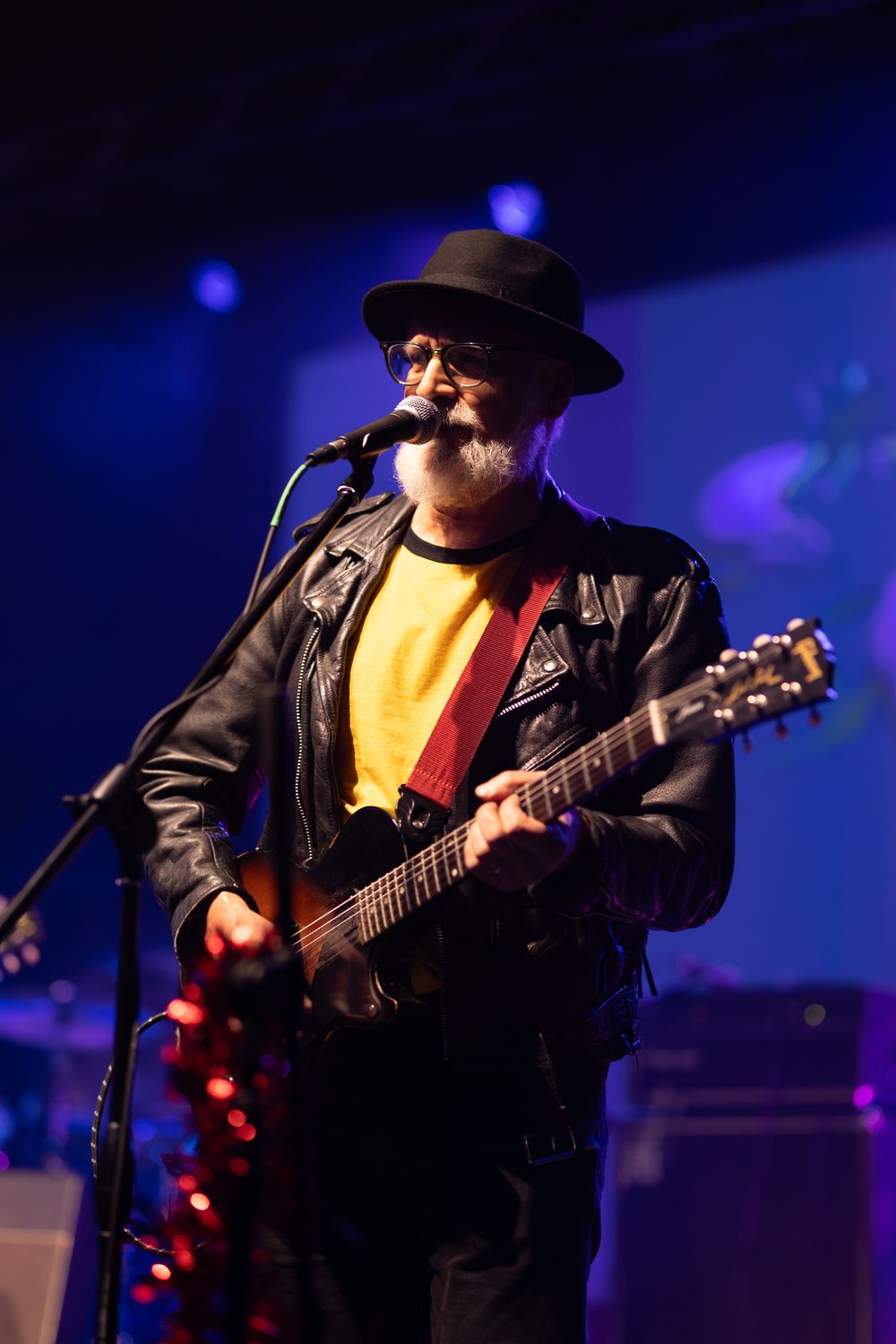 Older male musician with gray beard, black hat, and leather jacket playing acoustic guitar and singing into microphone on stage.