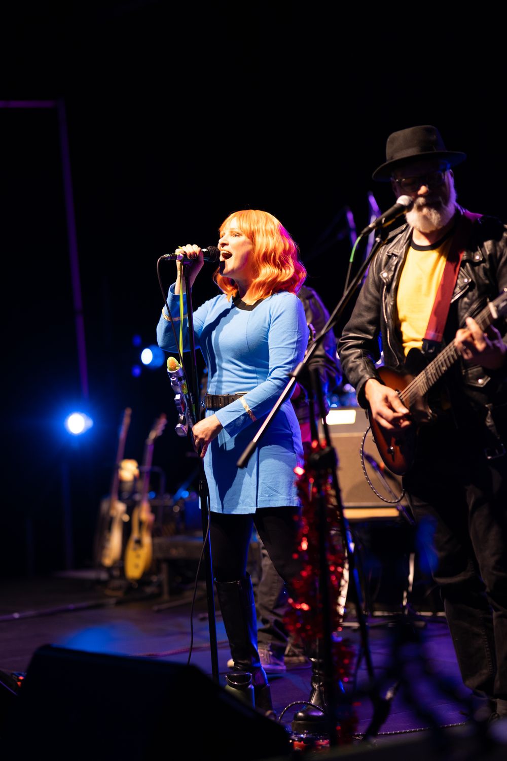 Female singer with red hair in blue dress performs on stage beside male guitarist in black hat and leather jacket.