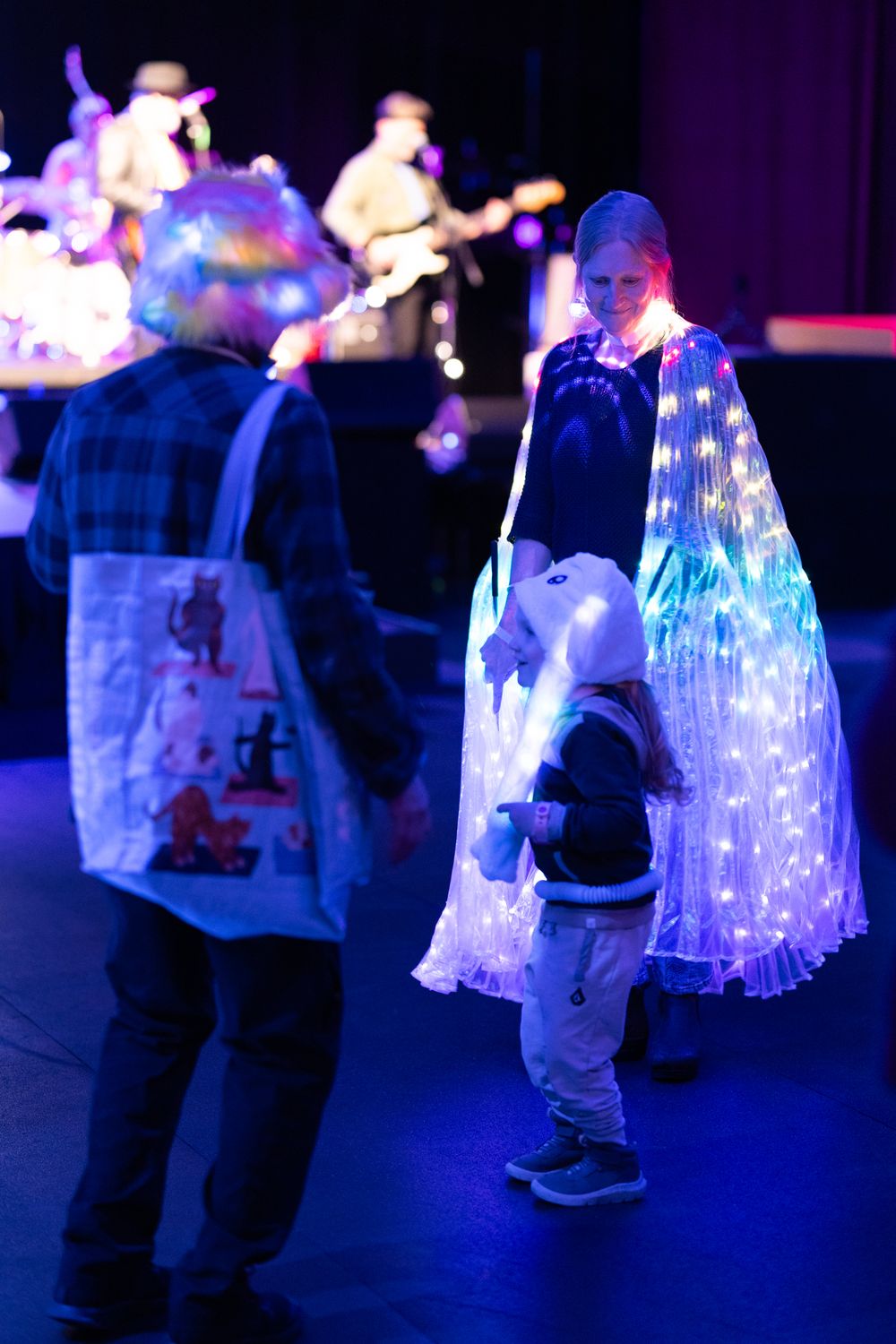 Woman in glowing LED cape stands with child at a live concert venue with band performing on stage in background.