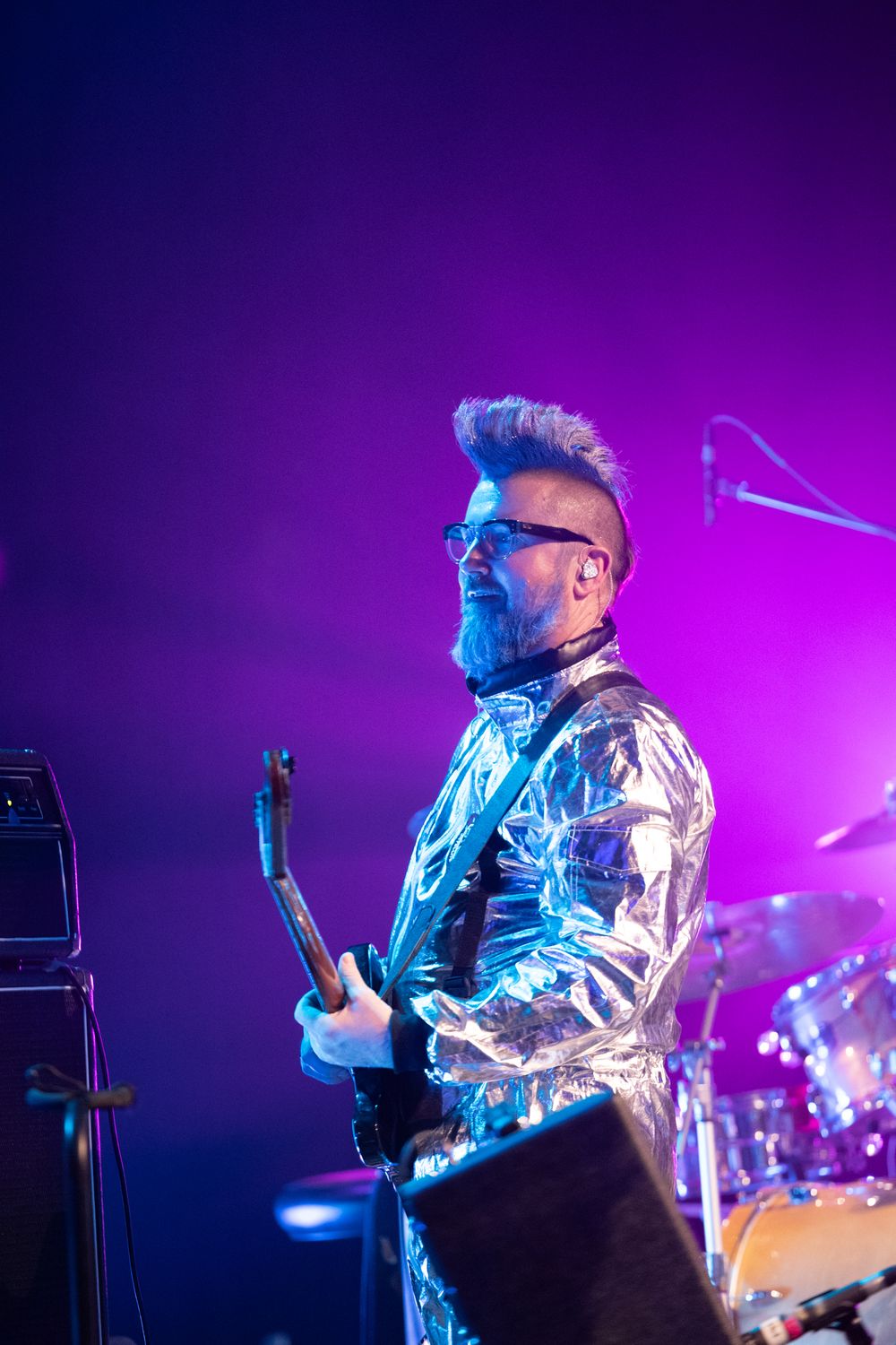 Male guitarist in silver metallic jacket plays electric guitar on stage under purple and blue concert lights.