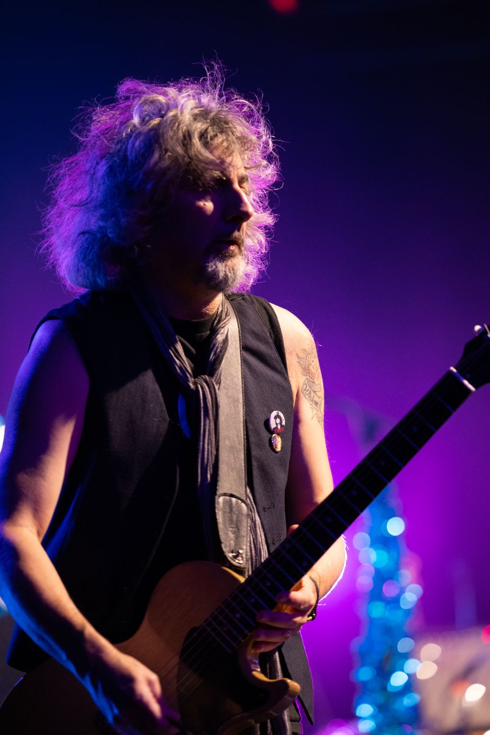 Male guitarist with wild curly hair plays electric guitar on stage under dramatic purple and blue stage lighting.