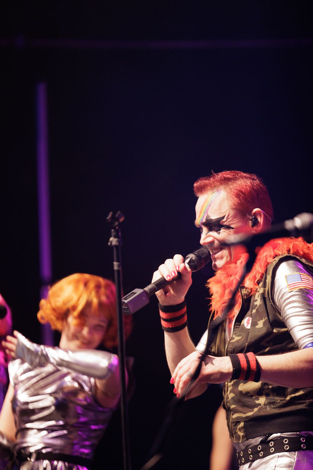 Red-haired punk rock singer performs on stage wearing camo vest with orange feather boa and dramatic face paint.