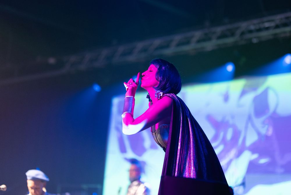 Female singer performs on stage in a flowing black cape under vibrant purple and pink stage lights.