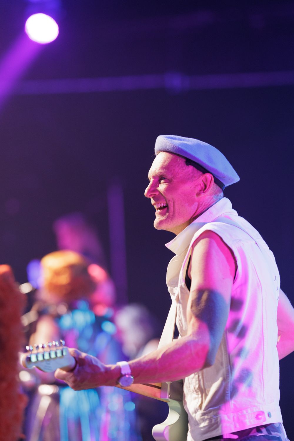 Smiling guitarist in white beret and vest performs on stage under colorful purple and pink concert lights.