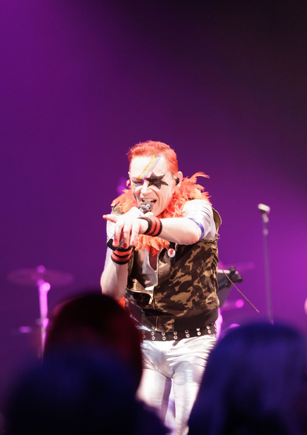Dramatic red-haired punk performer in camo vest points at audience under purple stage lighting.