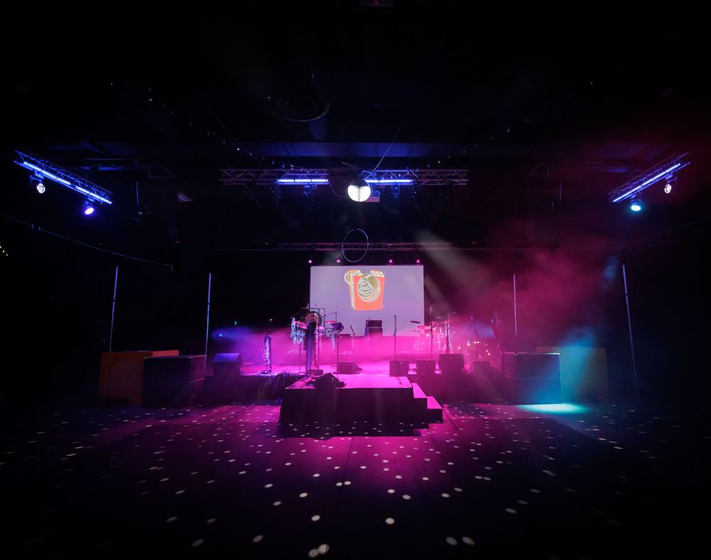 Empty event venue stage with purple and pink lighting and logo projected on screen.