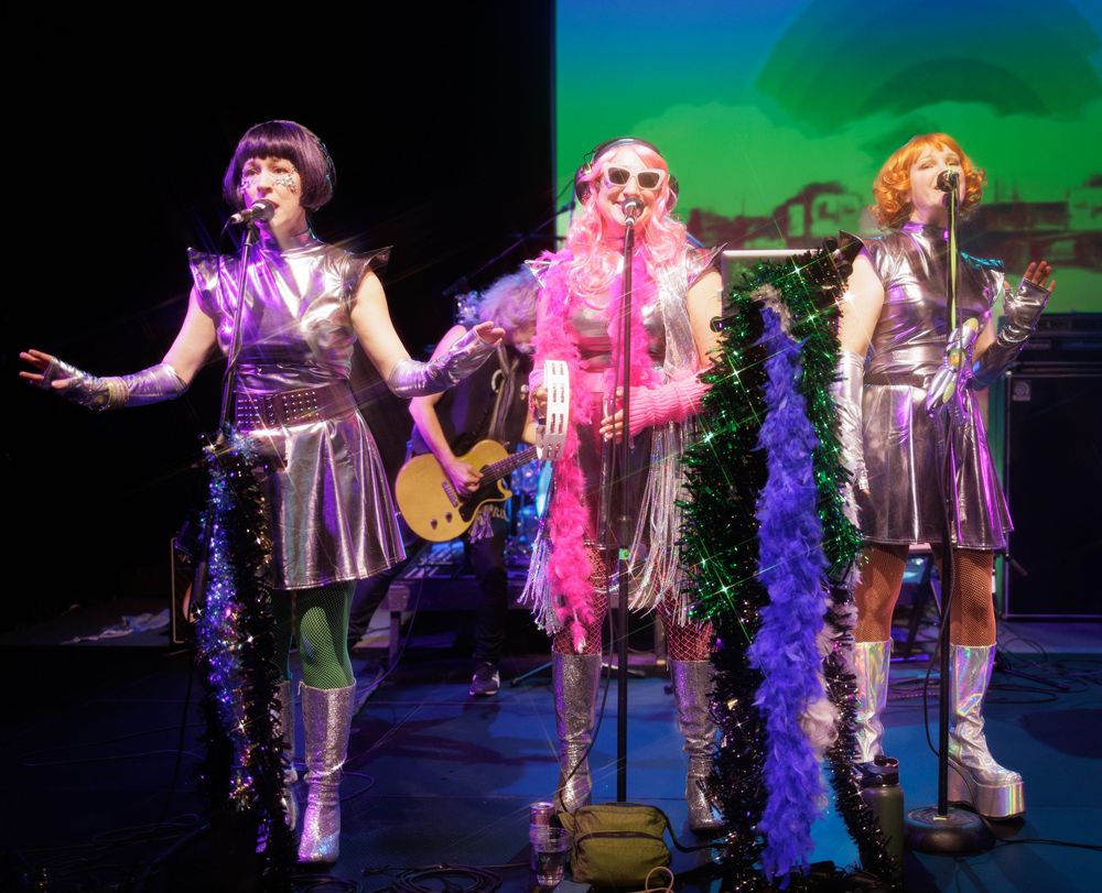 Three female performers in silver metallic costumes with colorful feather boas singing on a theatrical stage.