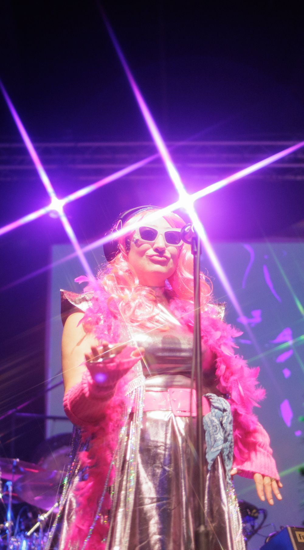 Female performer in silver dress and pink feather boa with sunglasses sings on stage under pink laser lights.