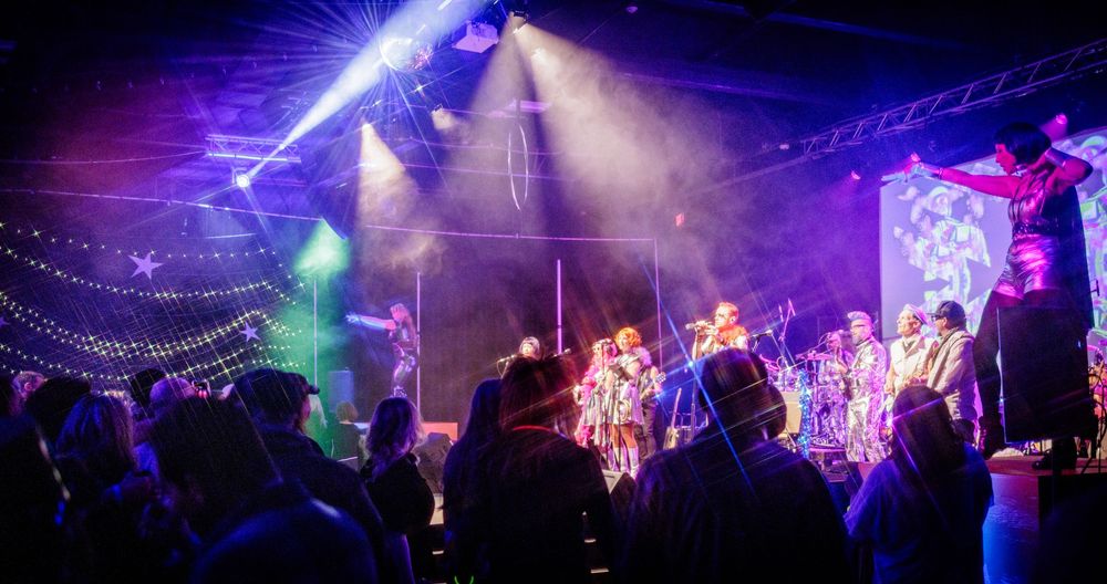 Large band performs on a colorfully lit stage before a crowd at an energetic live music concert event.