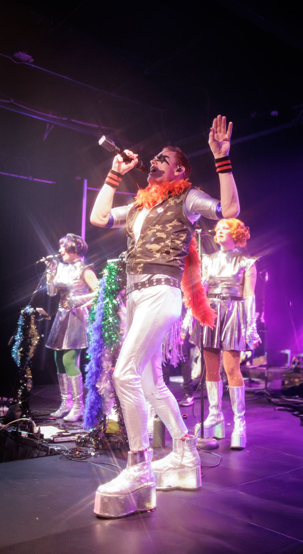 Flamboyant lead singer in platform boots and orange feather boa performs with backup dancers on colorful stage.