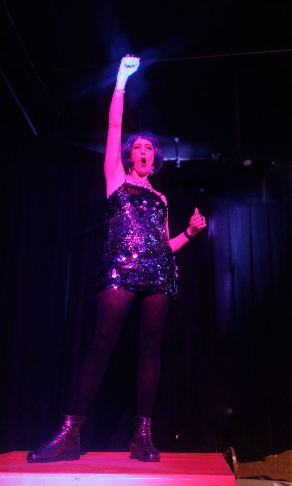 Energetic performer in sequined dress raises fist triumphantly under pink spotlight on dark stage.