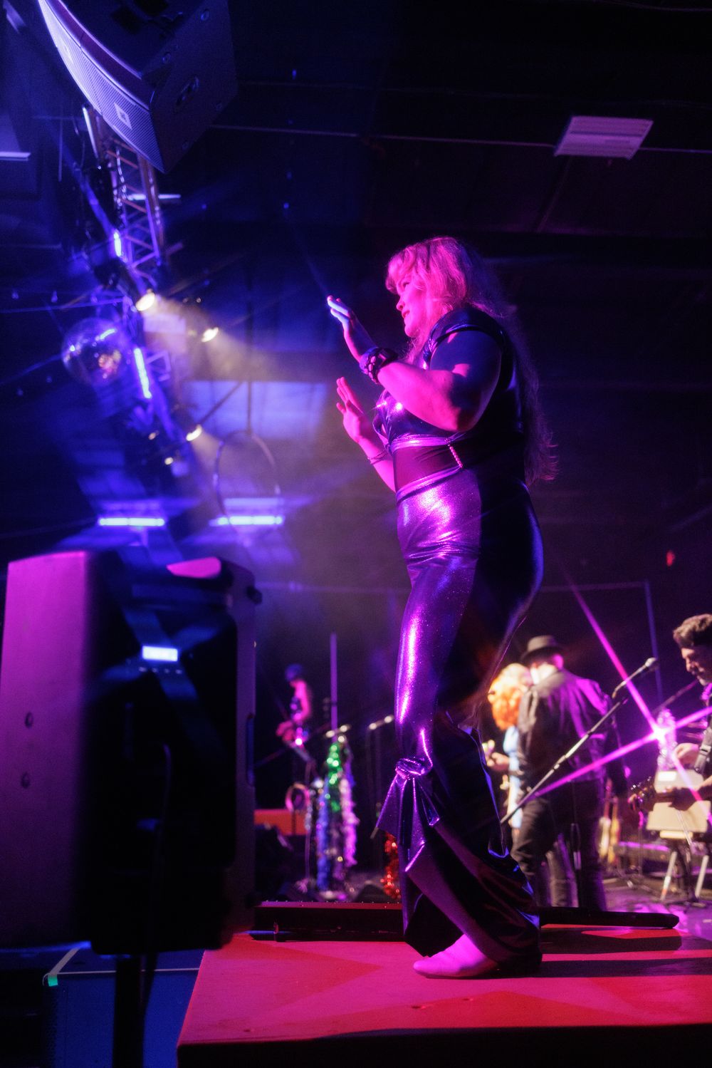 Female guitarist in metallic purple pants performs on stage under dramatic concert lighting with lens flares.