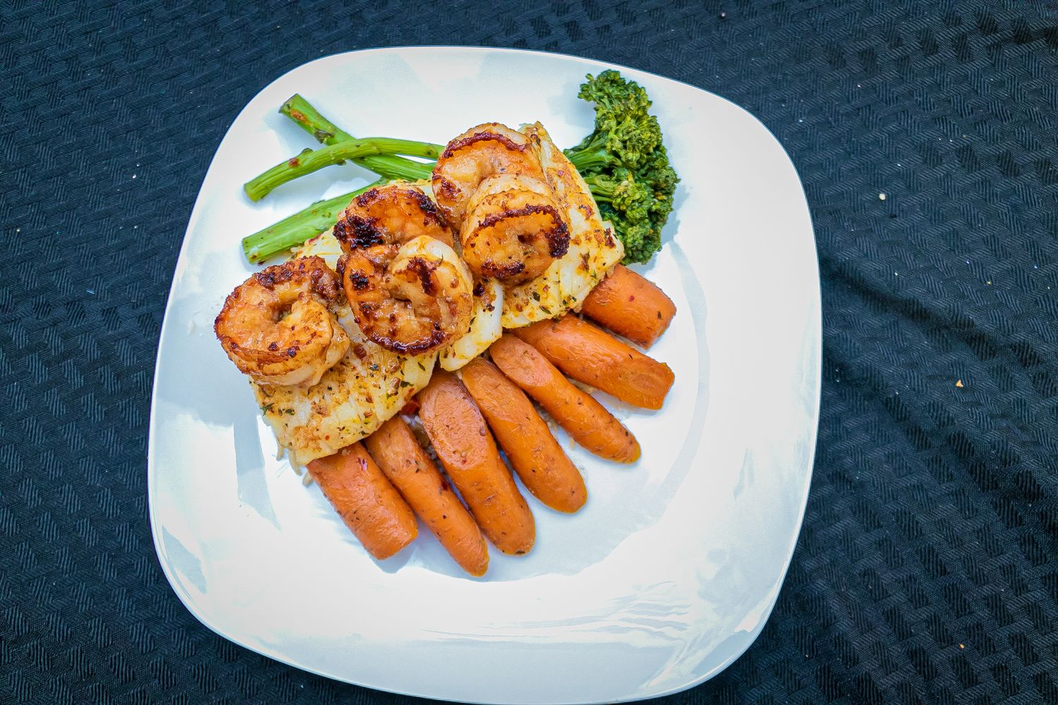Grilled shrimp and carrots served with green beans and broccoli on a white square plate against dark background.