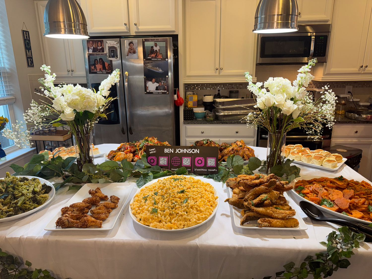 Catered buffet spread with various dishes, flowers, and appetizers on a white tablecloth.