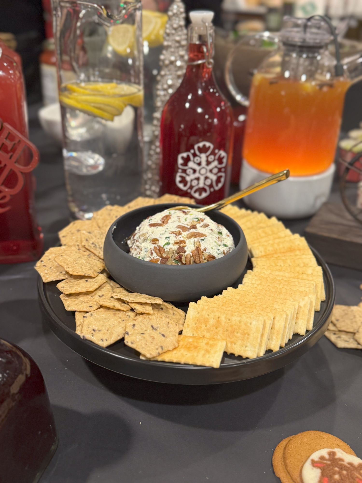 Appetizer platter with crackers and dip surrounded by festive holiday drinks and decorative glasses.