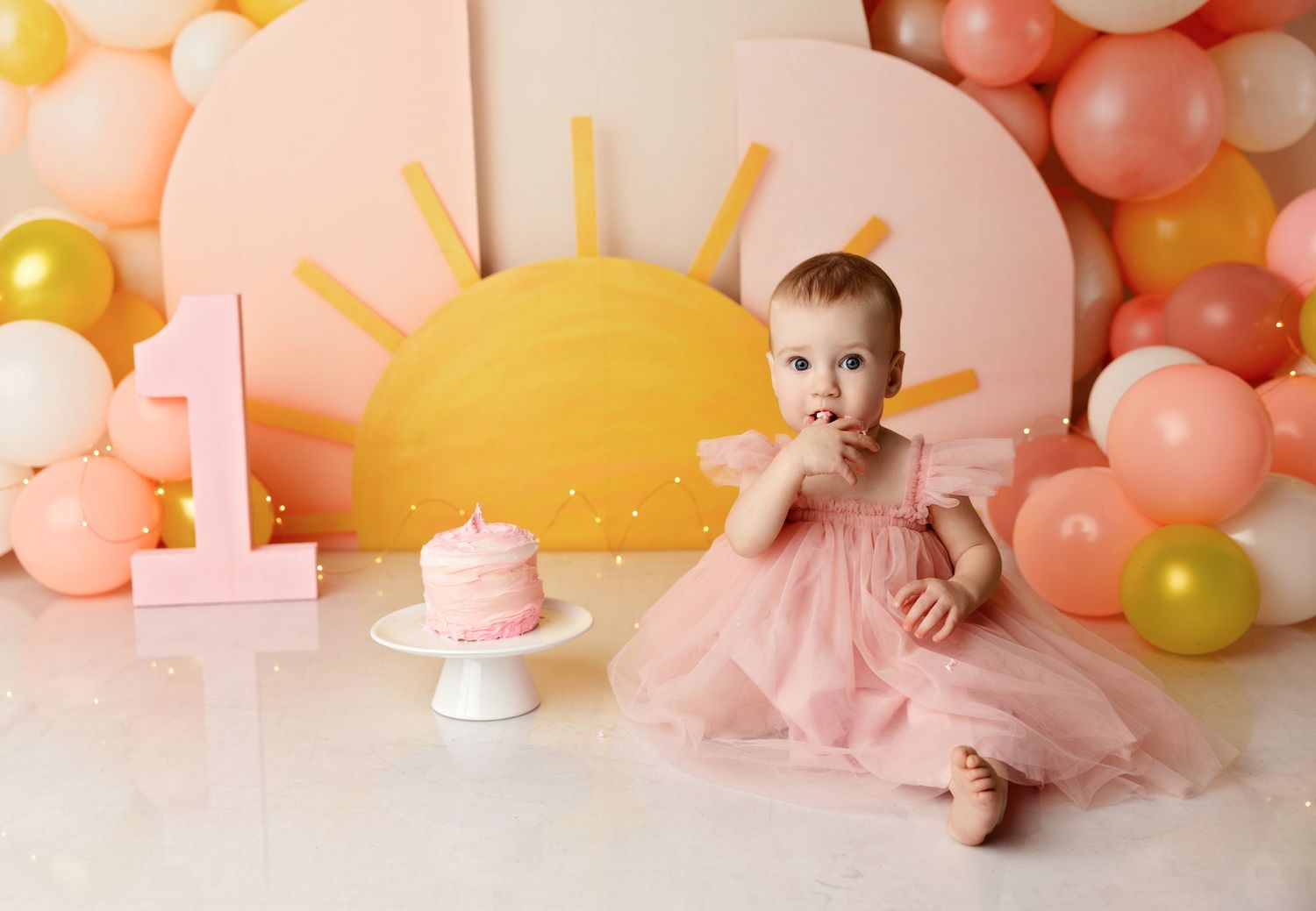 Tilton NH cake smash photography of a one-year-old girl in a pink dress, pastel balloon backdrop and first birthday studio portrait.