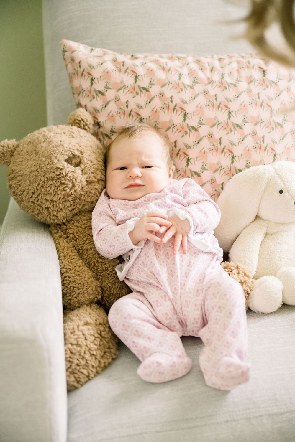 A sleeping baby in pink pajamas rests peacefully next to a plush teddy bear on a white couch.