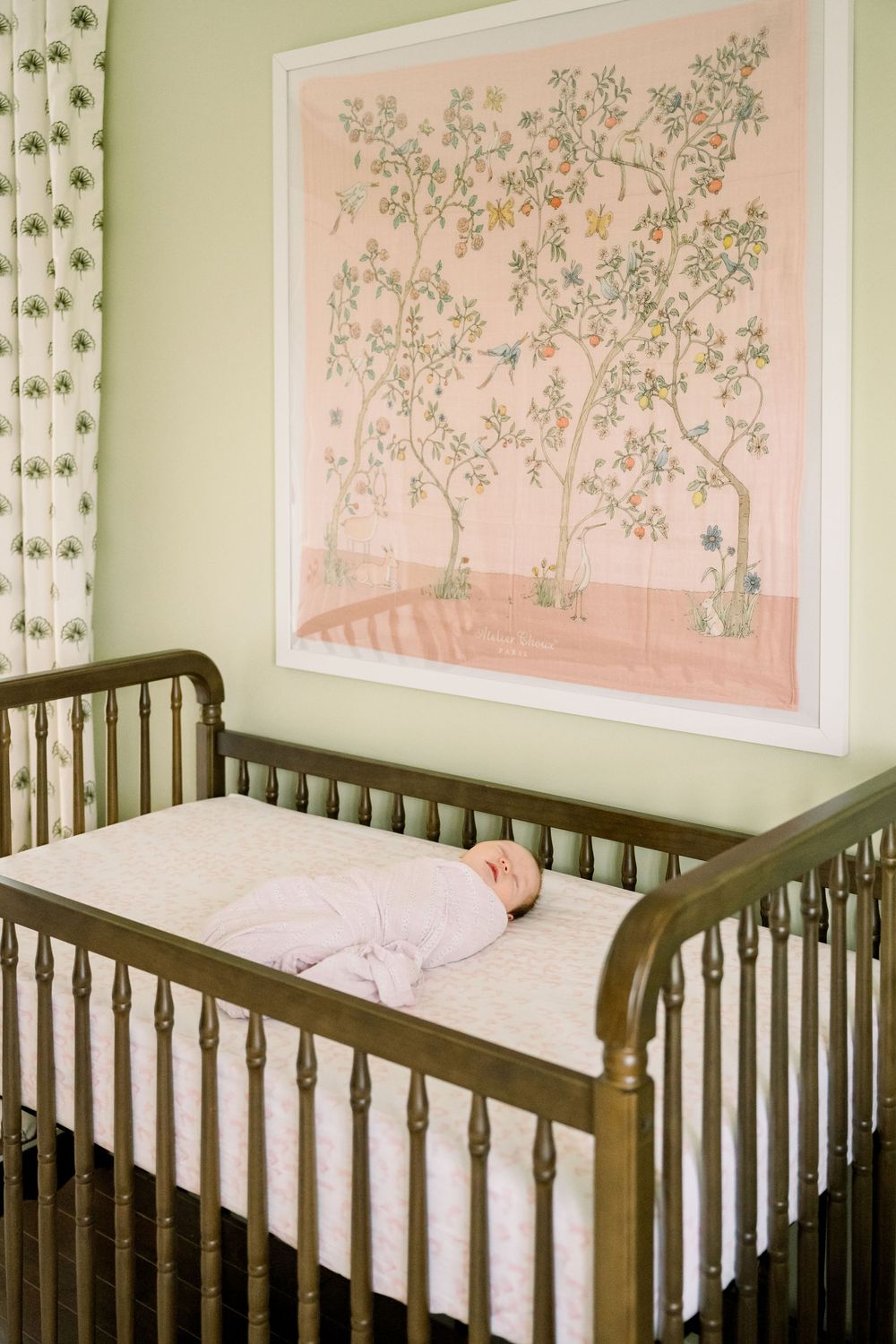 A wooden crib in a nursery with pink floral artwork on the wall and white bedding.