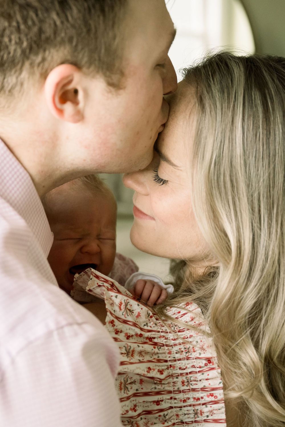 A tender family moment as parents share an intimate kiss while holding their sleeping newborn baby in a floral outfit.