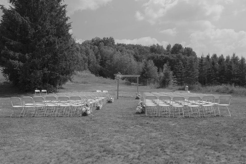 Black and white landscape view of white chairs arranged outdoors for a wedding ceremony with trees in background.