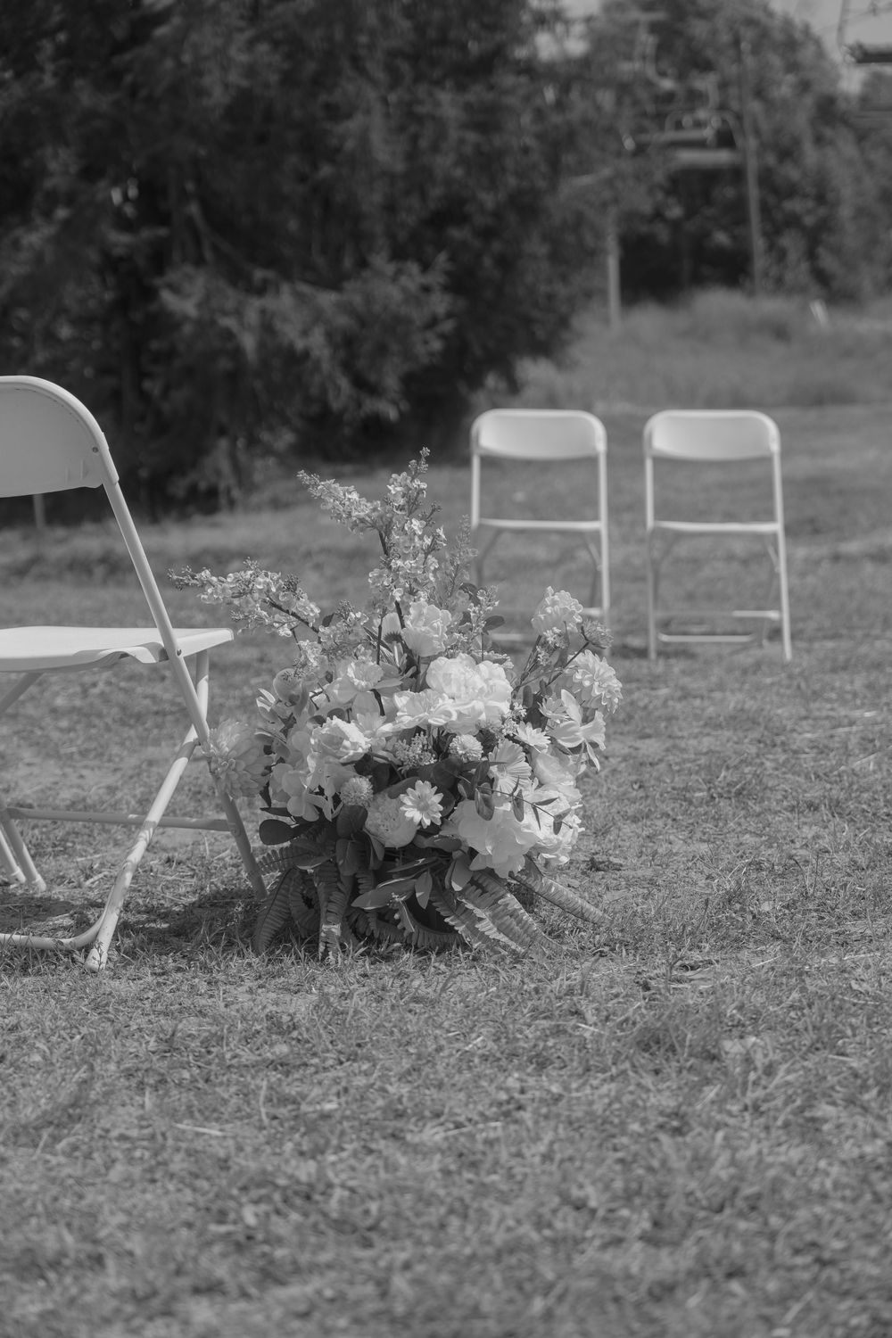 Black and white image of vintage white chairs arranged outdoors with floral decorations on the ground.
