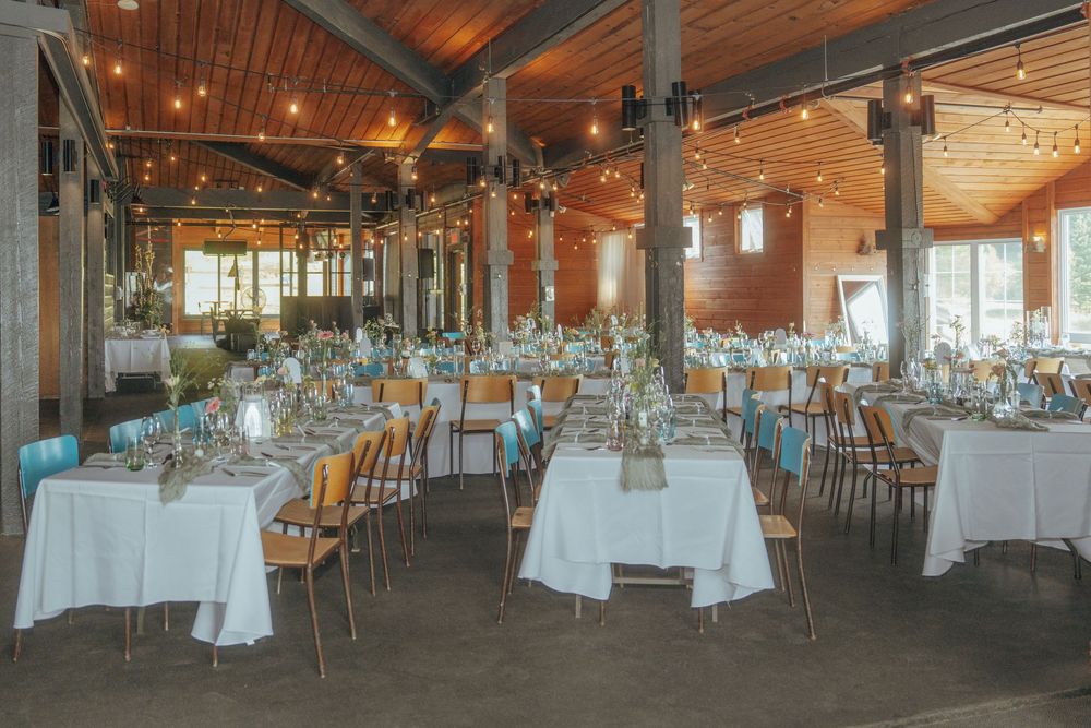 Large reception hall with wooden ceiling beams, white tablecloths, and ambient lighting setup for a wedding.