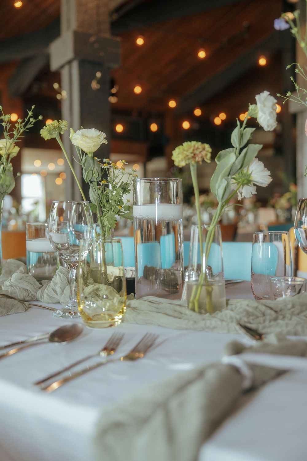 Wedding reception table settings with white flowers in glass vases, turquoise candles, and string lights overhead.