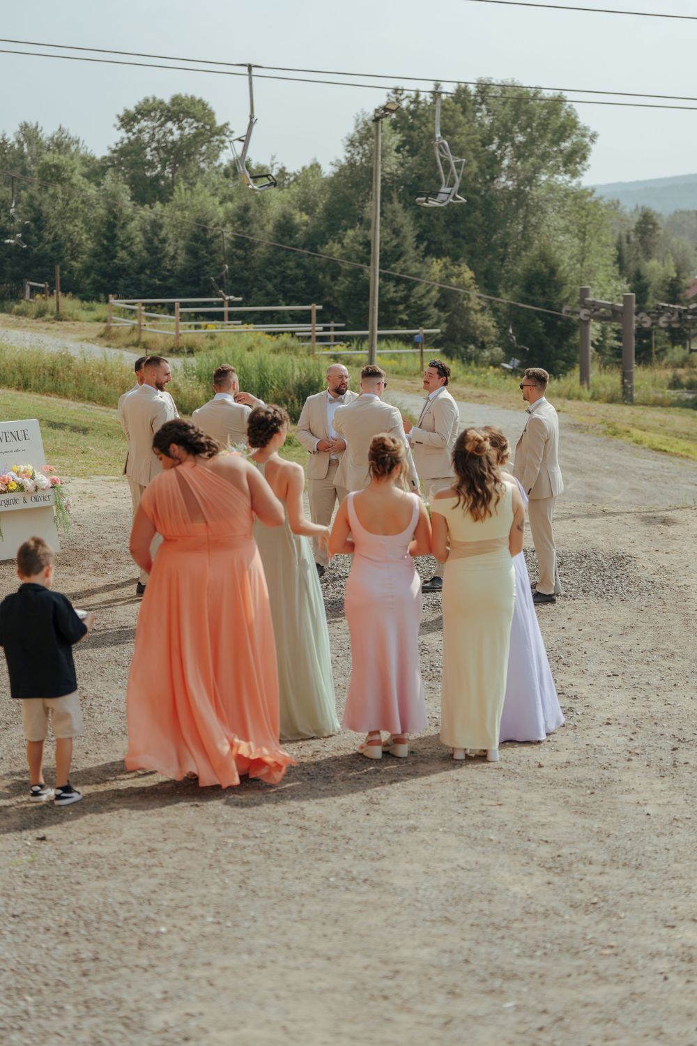 A wedding party in peach and white dresses poses together on a gravel path outdoors.