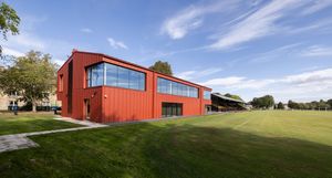 Modern red shipping container building with large windows sits on a grassy hill under blue skies with wispy clouds.