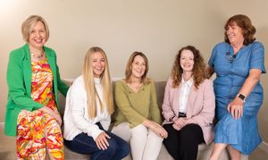 Five people sit together on a beige couch wearing colorful outfits including green, white, olive, pink and blue clothing.