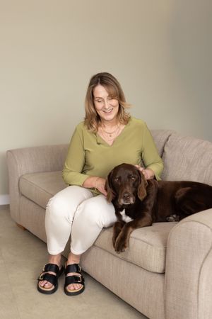 A chocolate Labrador retriever lounges comfortably on a beige couch next to someone wearing a green sweater.