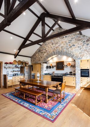 Rustic dining room with exposed wooden beams, stone walls, hardwood floors and a colorful oriental rug under a long table.
