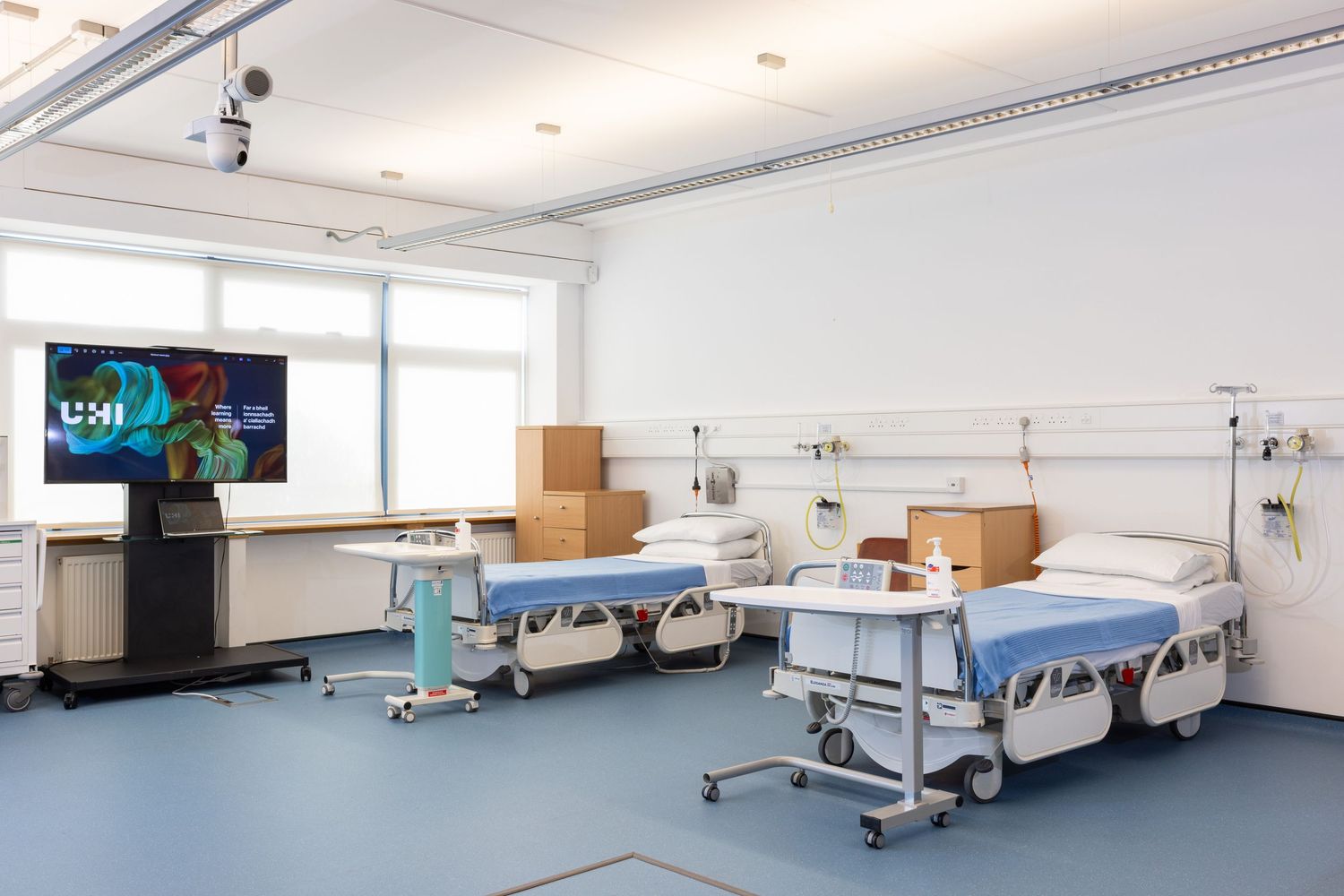 Modern hospital room with multiple patient beds, medical equipment, and large windows providing natural light.