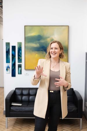 Professional woman in a beige blazer gestures animatedly while standing in front of modern artwork and leather couch.