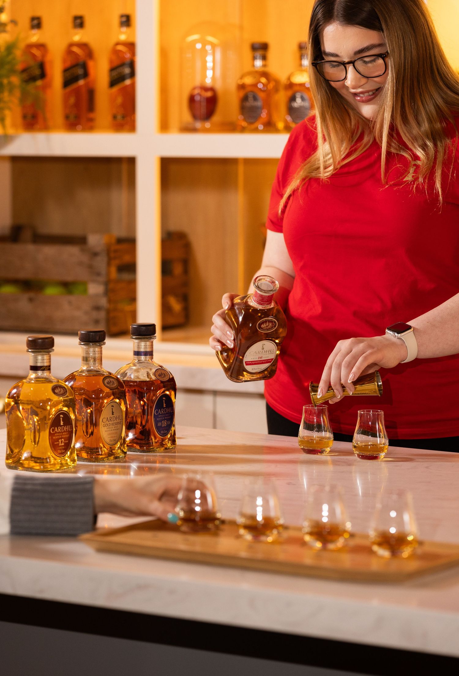 A bartender in red pours whiskey samples for a tasting flight with multiple bottles displayed on shelves.