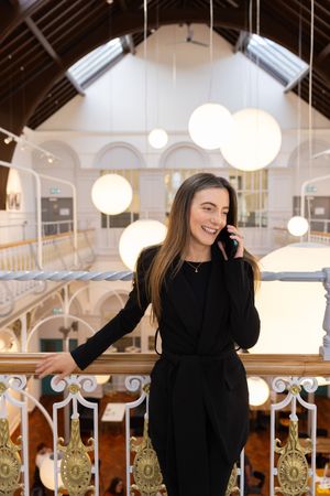 A woman in a black sweater smiles and poses in a bright interior space with hanging pendant lights and ornate decor.